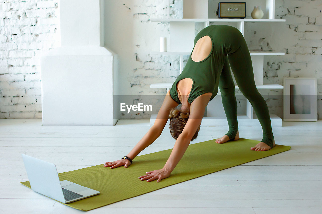 A slender woman in a green sports jumpsuit performs yoga exercise at home, in a light interior,