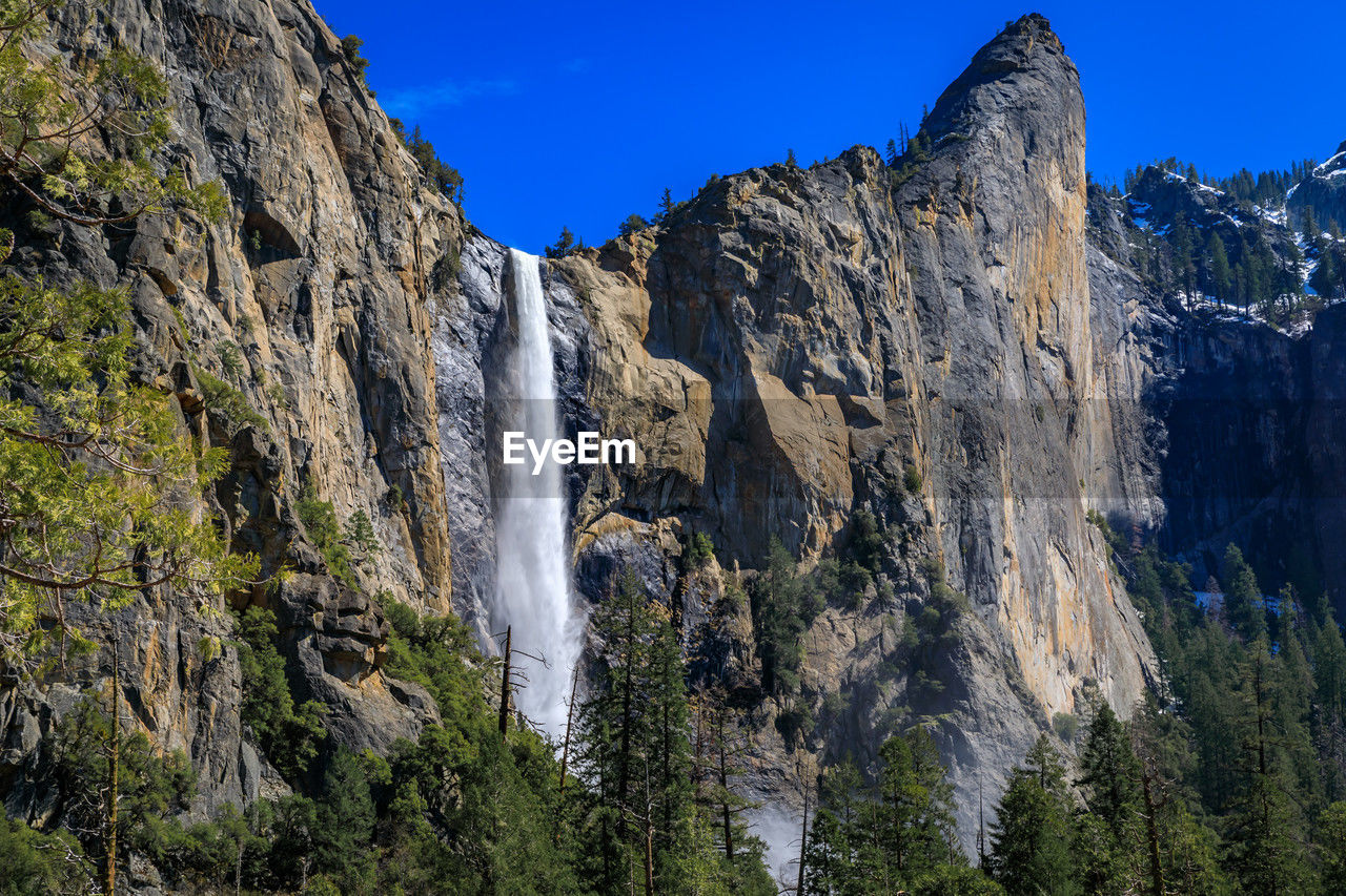 waterfall, scenics - nature, beauty in nature, rock, mountain, environment, nature, land, national park, plant, wilderness, water, sky, landscape, tree, travel destinations, cliff, rock formation, mountain range, travel, park, adventure, forest, non-urban scene, blue, terrain, no people, tourism, outdoors, motion, pine tree, coniferous tree, mountain peak, ridge, steep, clear sky, activity, formation, cold temperature, geology, walking, water feature, pinaceae, pine woodland, idyllic, snow, long exposure, tranquil scene, day, leisure activity, massif