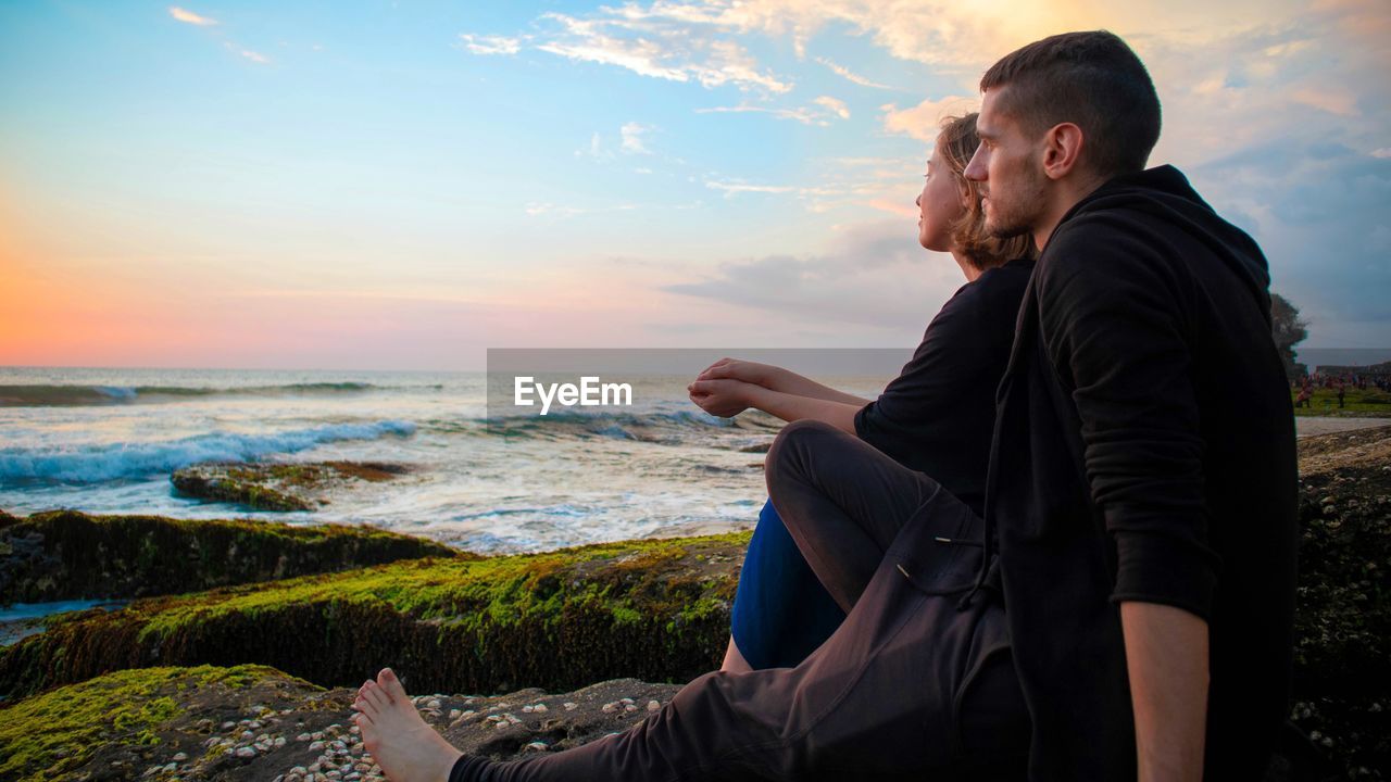 Side view of couple sitting on rock at beach against sky