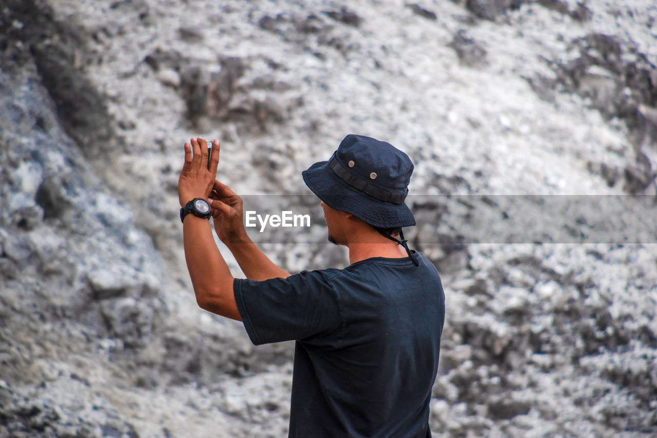 An asian man taking a picture with his cellphone on a rock, rear view 