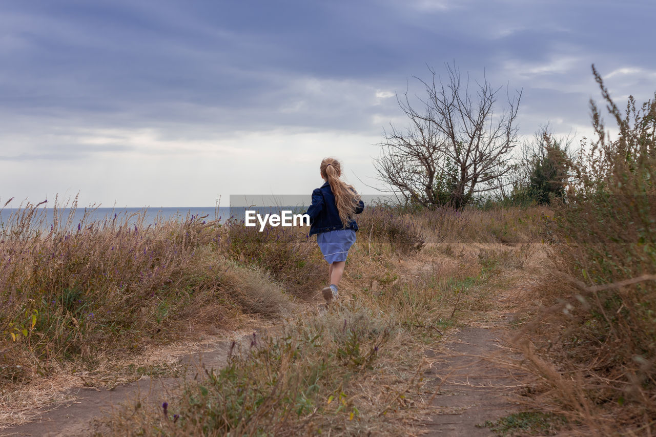 Blond long hair child girl in denim jacket walks on sea landscape. travelling hiking running outdoor