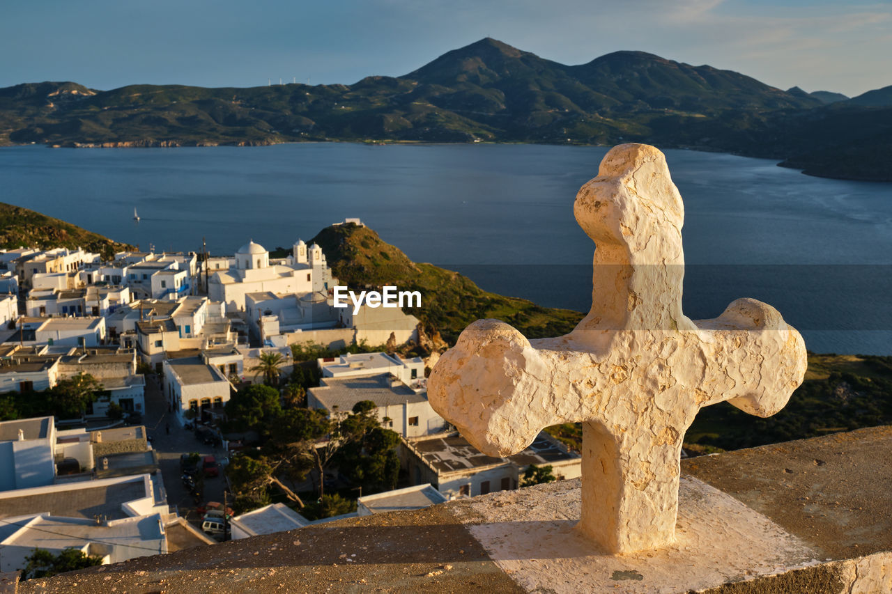 Christian cross and plaka village on milos island over red geranium flowers on sunset in greece