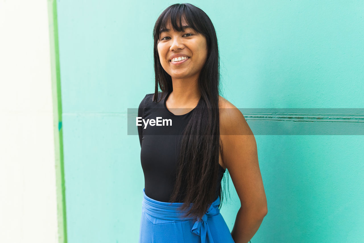 Cheerful young ethnic female with long hair smiling and looking at camera while standing against turquoise wall on sunny day on city street