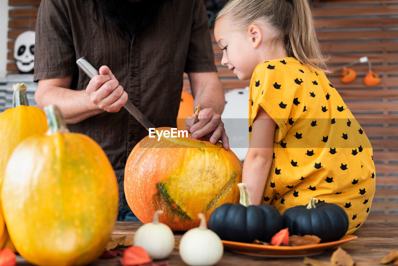 Midsection of father cutting pumpkin with girl sitting a home