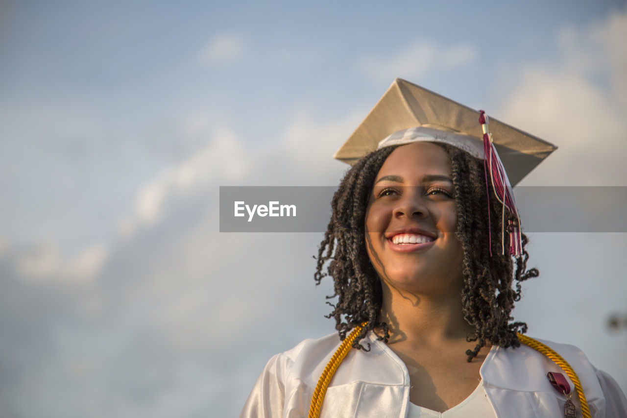 Smiling young woman wearing mortarboard against sky