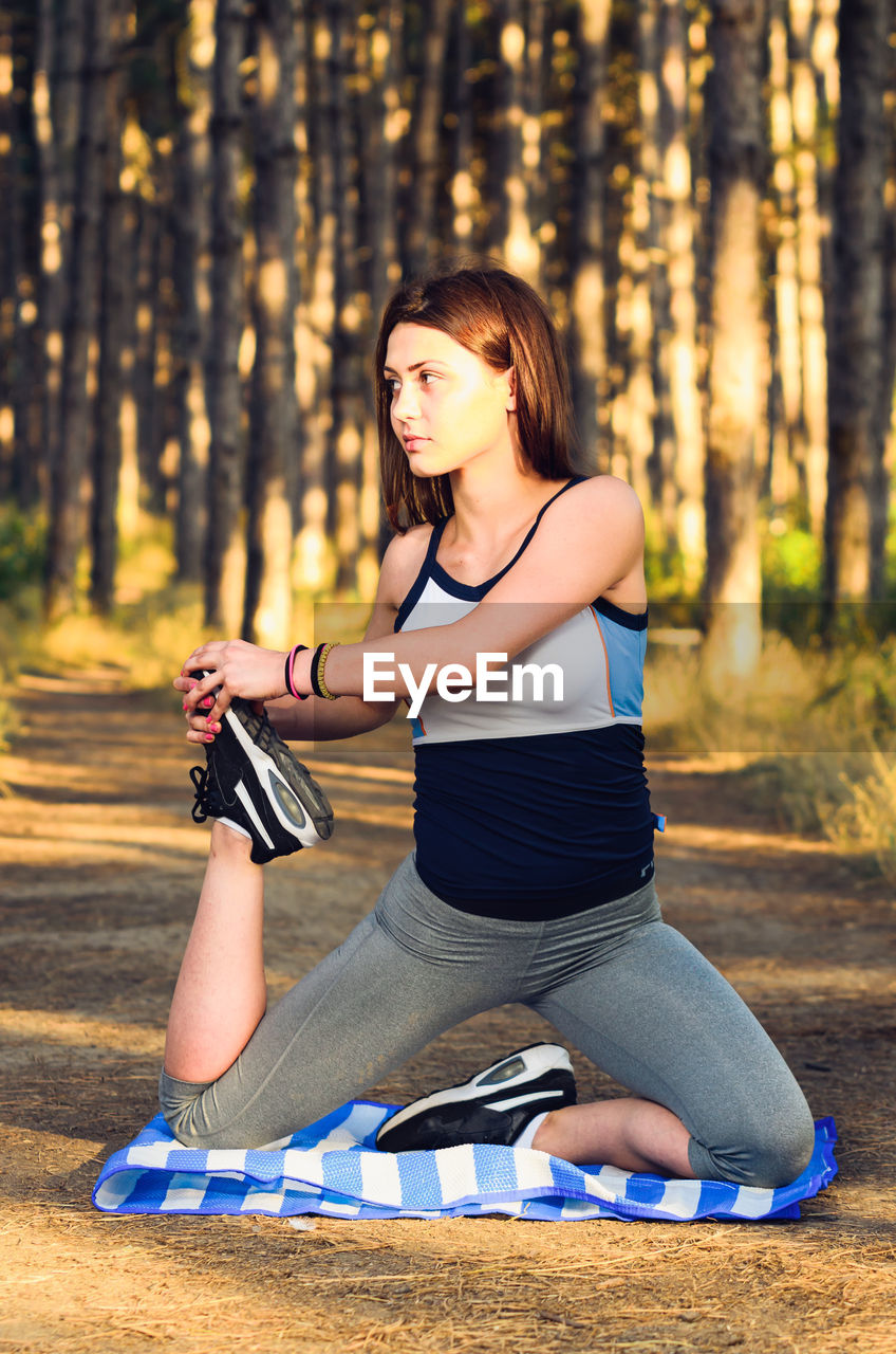 Young woman stretching body on exercise mat against trees in forest during sunset