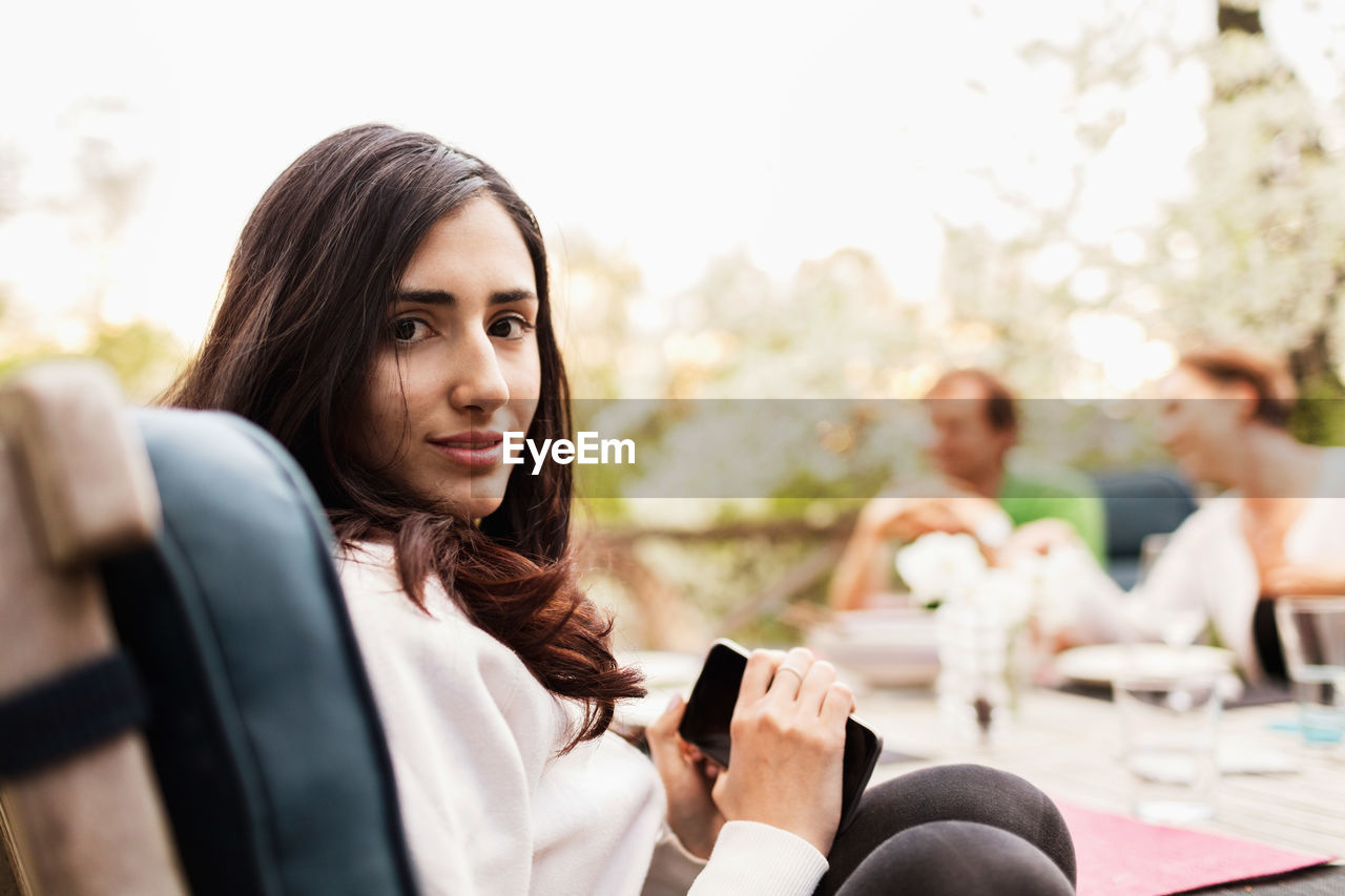 Side view portrait of teenage girl holding smart phone while parents sitting in background at yard