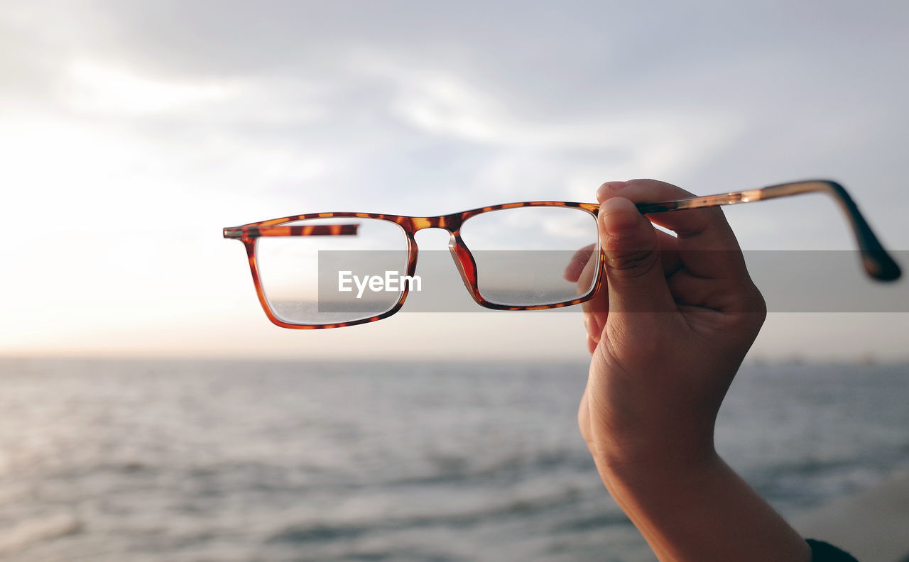 Close-up of hand holding eyeglasses against sky at beach