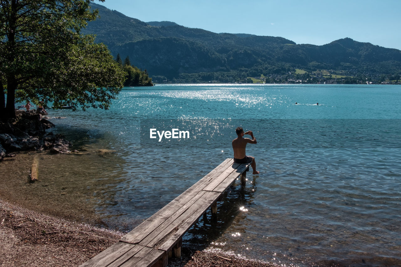 Rear view of man drinking beer while sitting on pier over lake