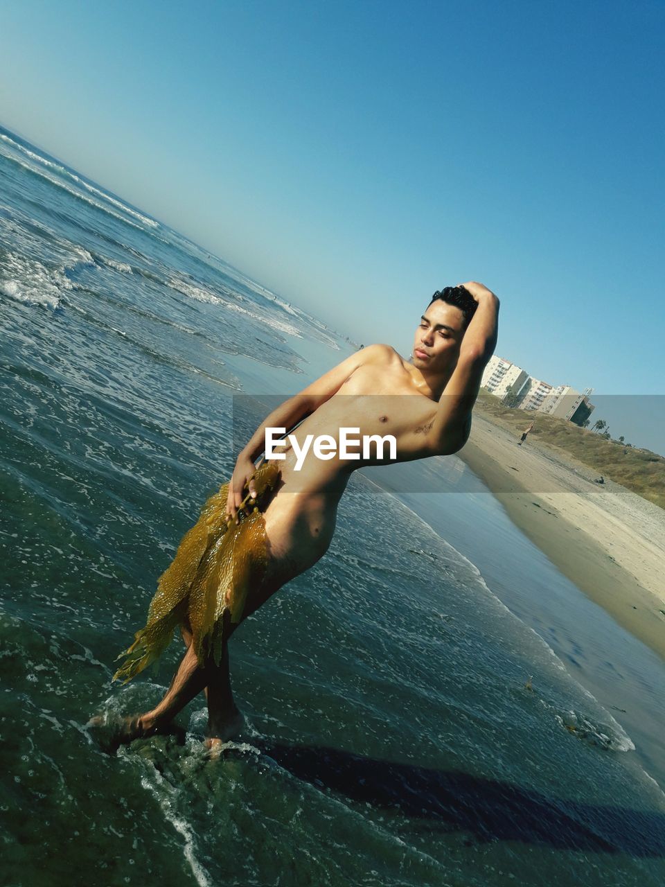 Naked young man posing at beach against clear sky