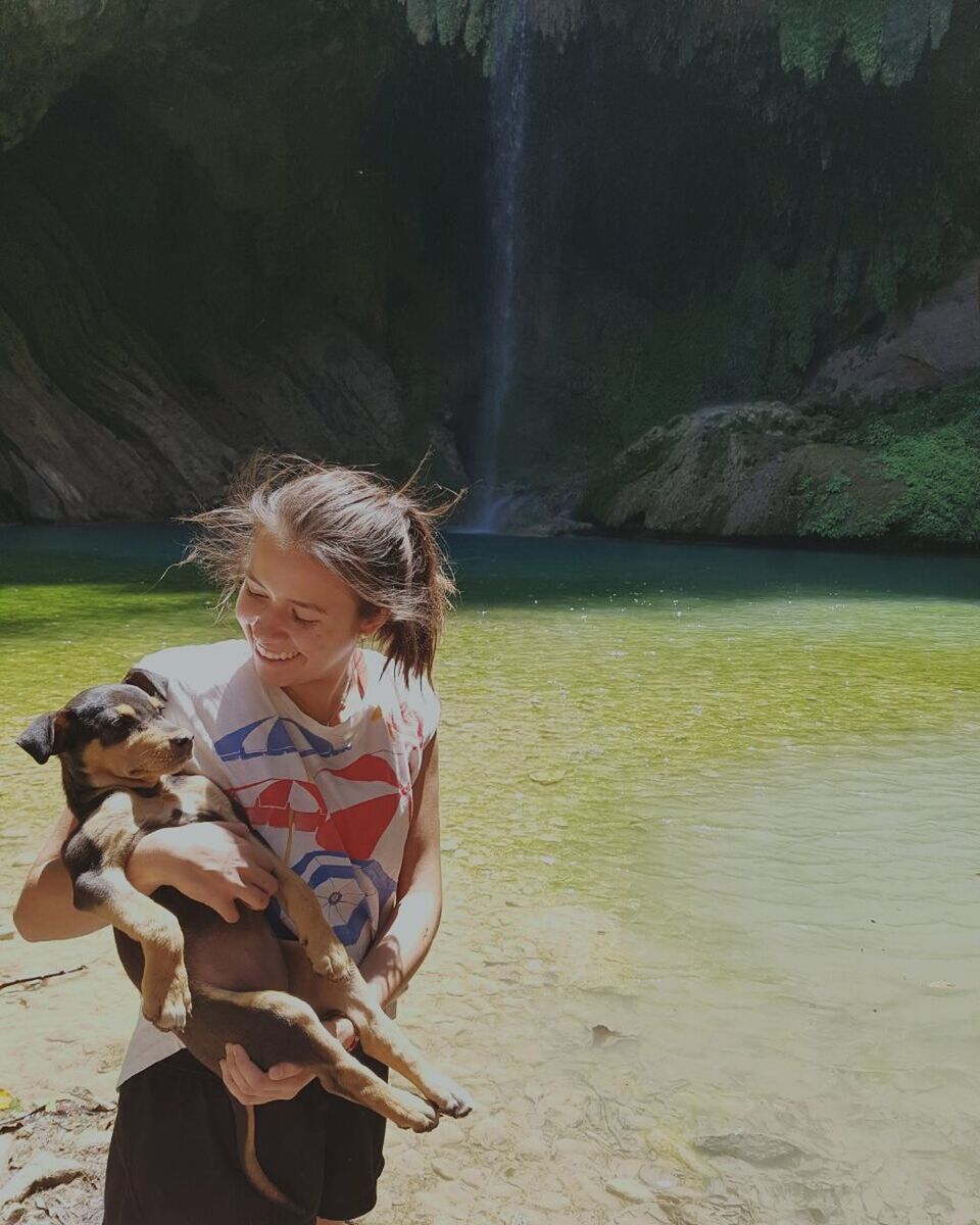 Smiling teenage girl holding puppy while standing against waterfall