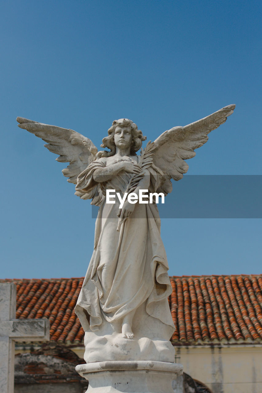 Cemetery statue. angel with wings. red tile roof in background.