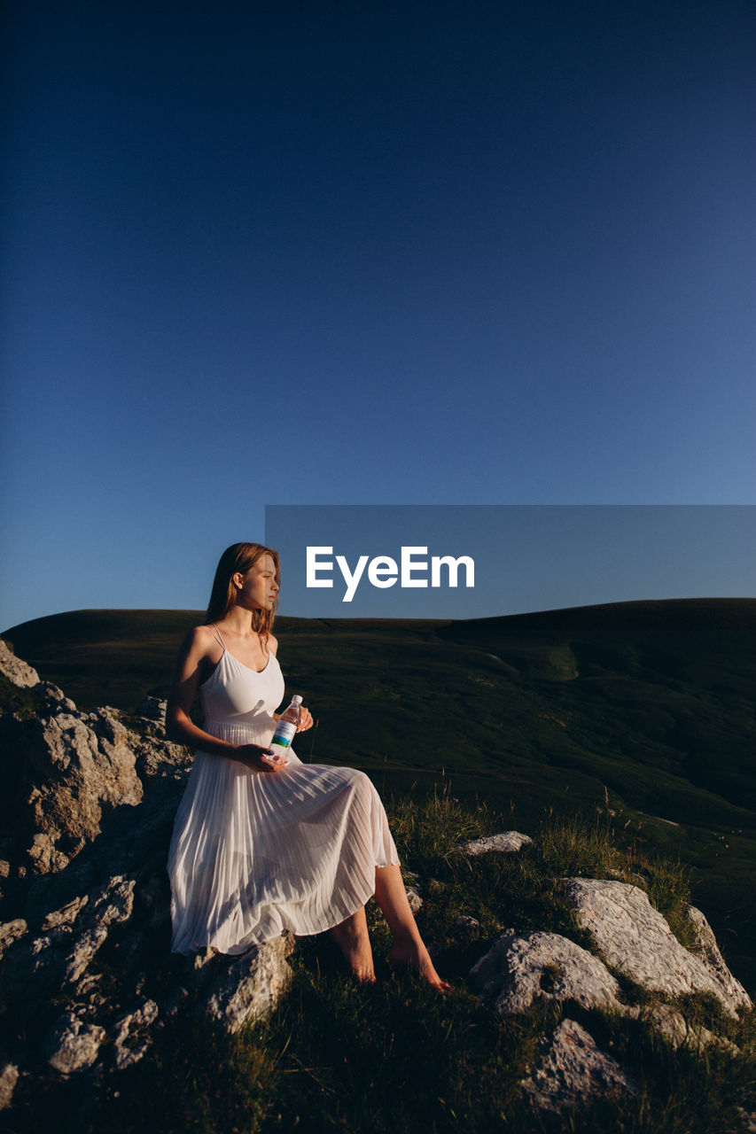 WOMAN SITTING ON ROCK AGAINST SKY