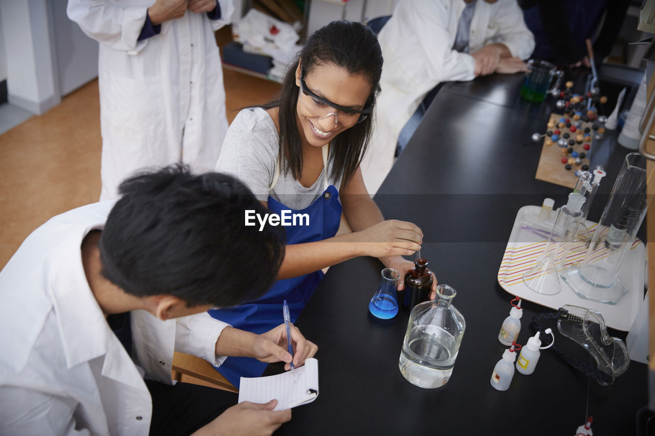 High angle view of smiling multi-ethnic students mixing solutions and writing in spiral notepad at chemistry laboratory