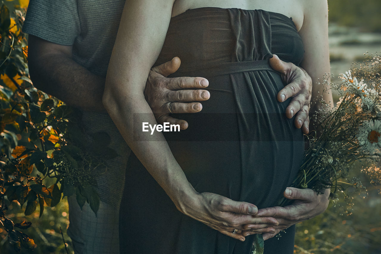 Pregnant woman and her husband hugging her tummy standing outdoors surrounded by nature