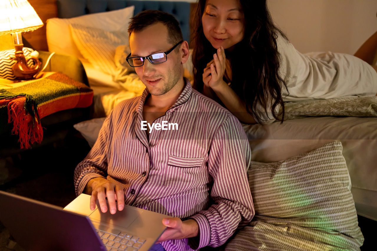 Couple watching funny content on laptop while resting on bed at home in evening.