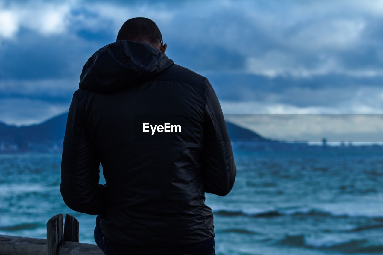 Rear view of man standing by sea against cloudy sky