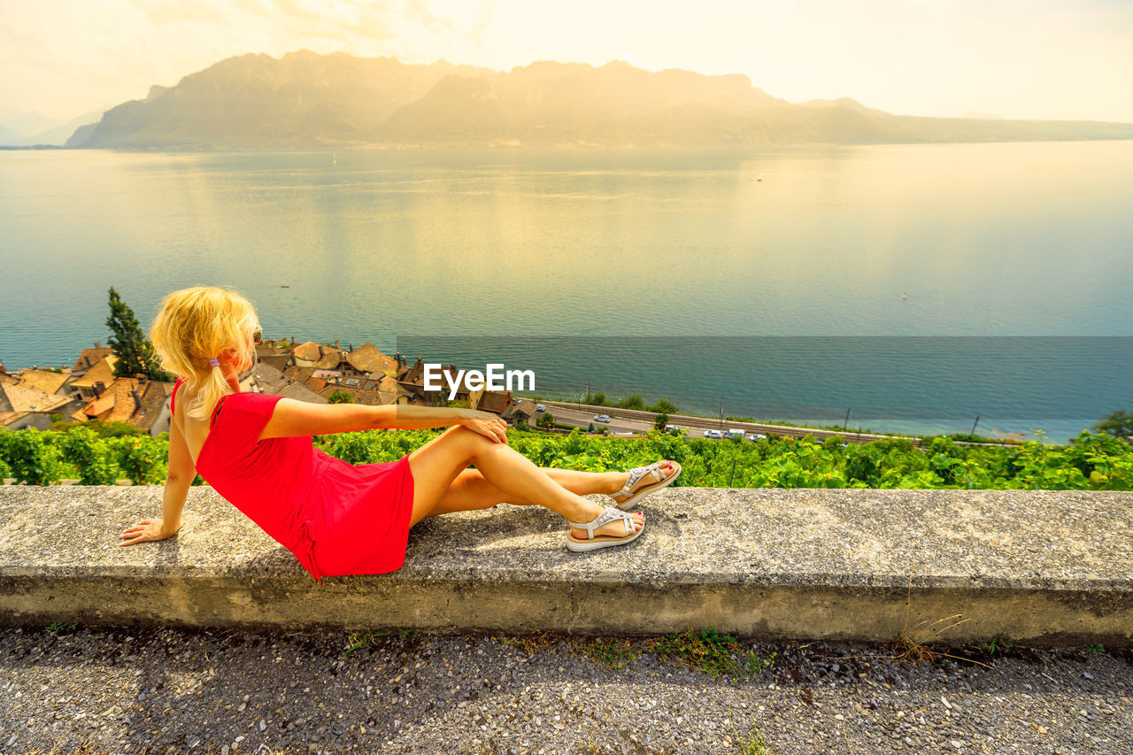 REAR VIEW OF WOMAN SITTING BY LAKE AGAINST MOUNTAINS