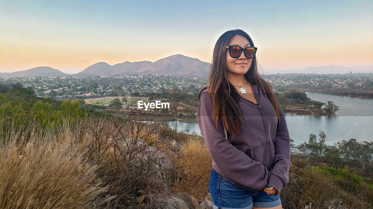 PORTRAIT OF SMILING YOUNG WOMAN STANDING AGAINST MOUNTAIN