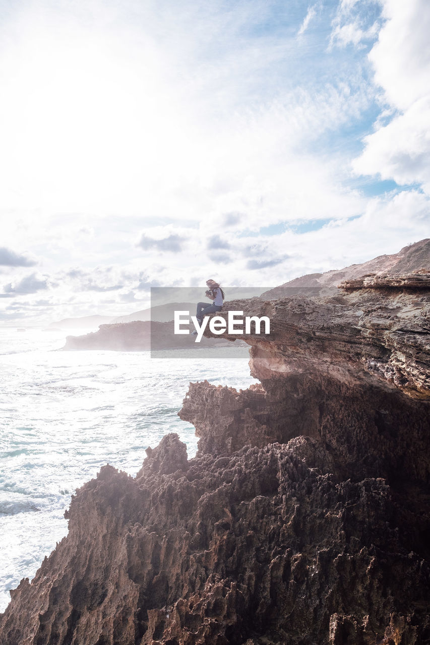 Woman sitting on cliff by sea against cloudy sky during sunny day