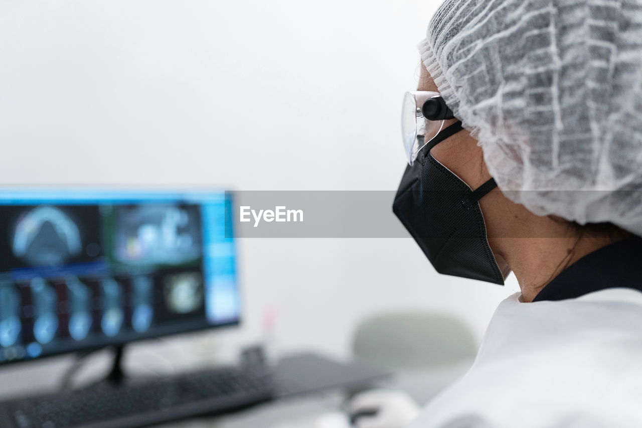 Side view of female medical specialist in protective mask and goggles working with scan image on computer monitor in modern clinic