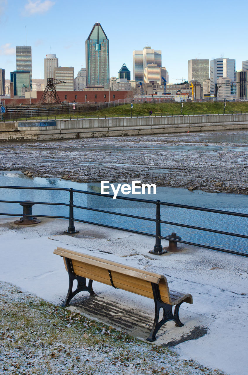 Empty bench by river in city against sky