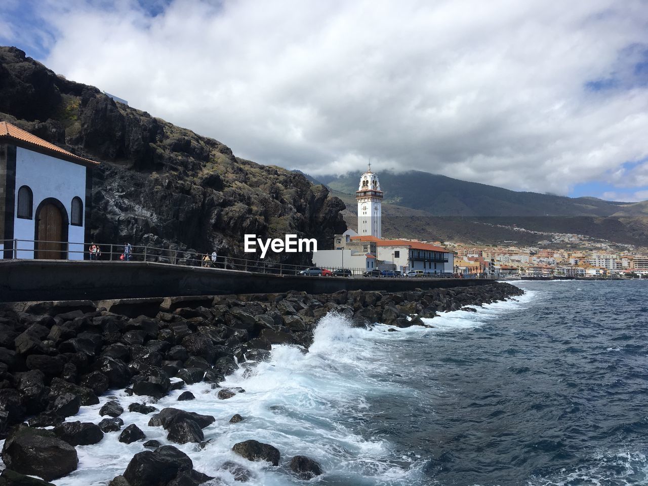 LIGHTHOUSE BY SEA AGAINST BUILDINGS AND MOUNTAIN