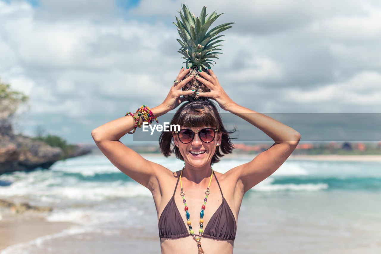 Portrait of seductive women holding pineapple at beach against sky