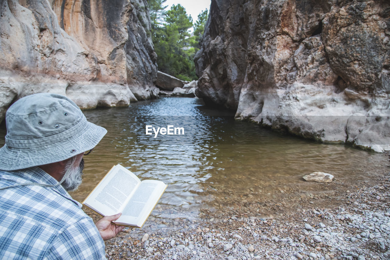 Tourist with a beard and hat reads a book sitting on the river bank