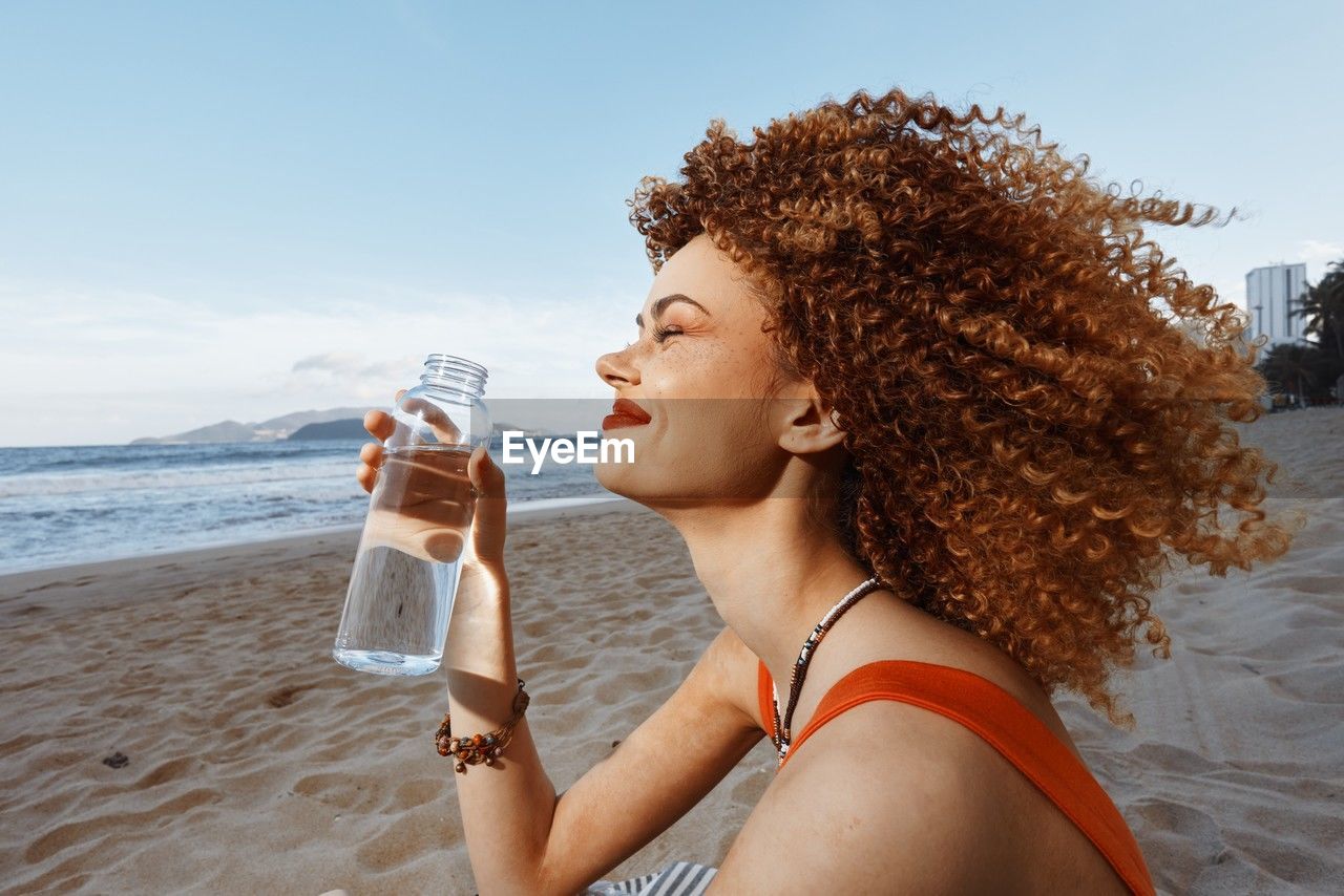 side view of woman drinking water while sitting on beach against sky