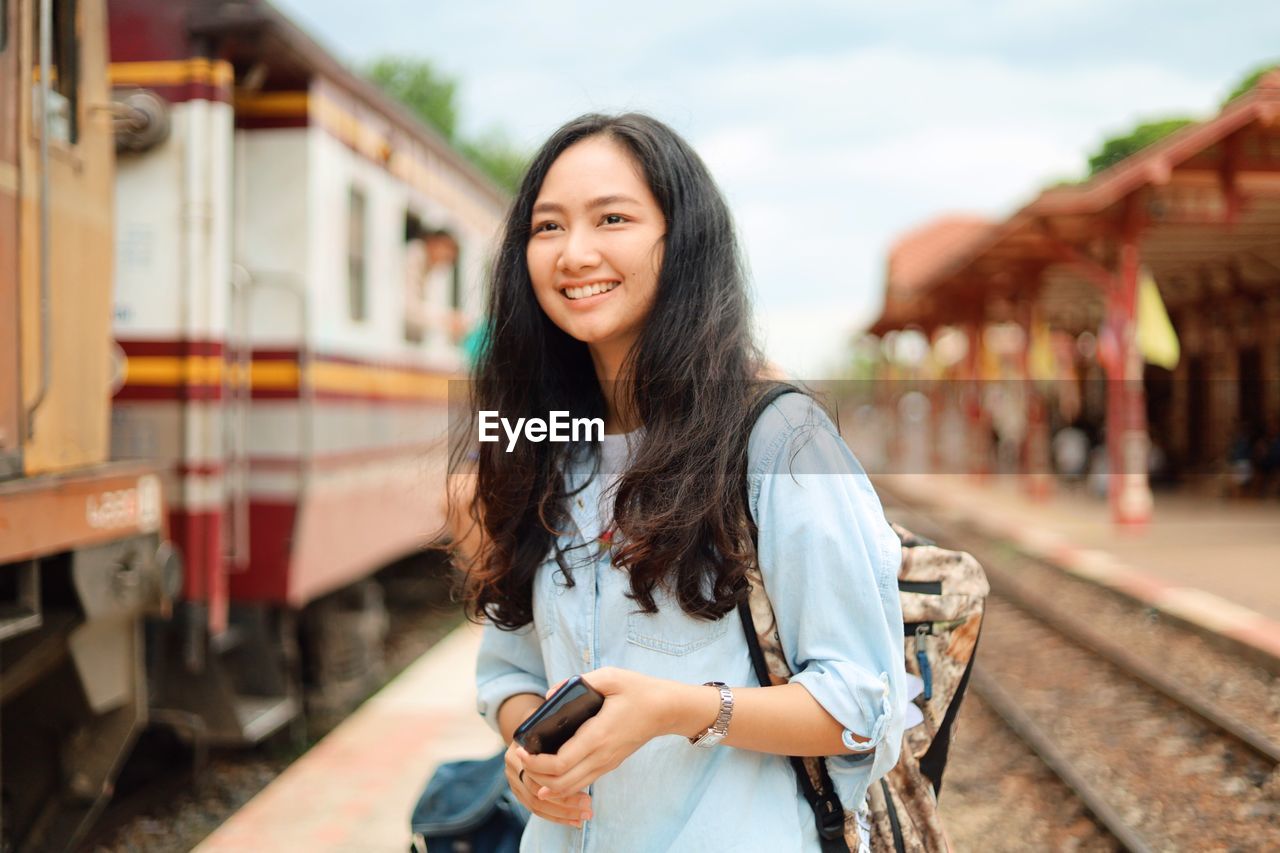 PORTRAIT OF SMILING YOUNG WOMAN STANDING ON RAILROAD TRACKS