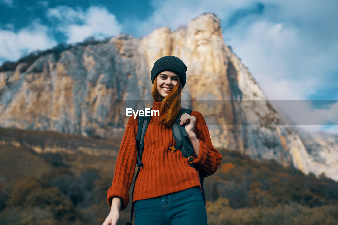 YOUNG WOMAN WEARING HAT STANDING AGAINST MOUNTAIN