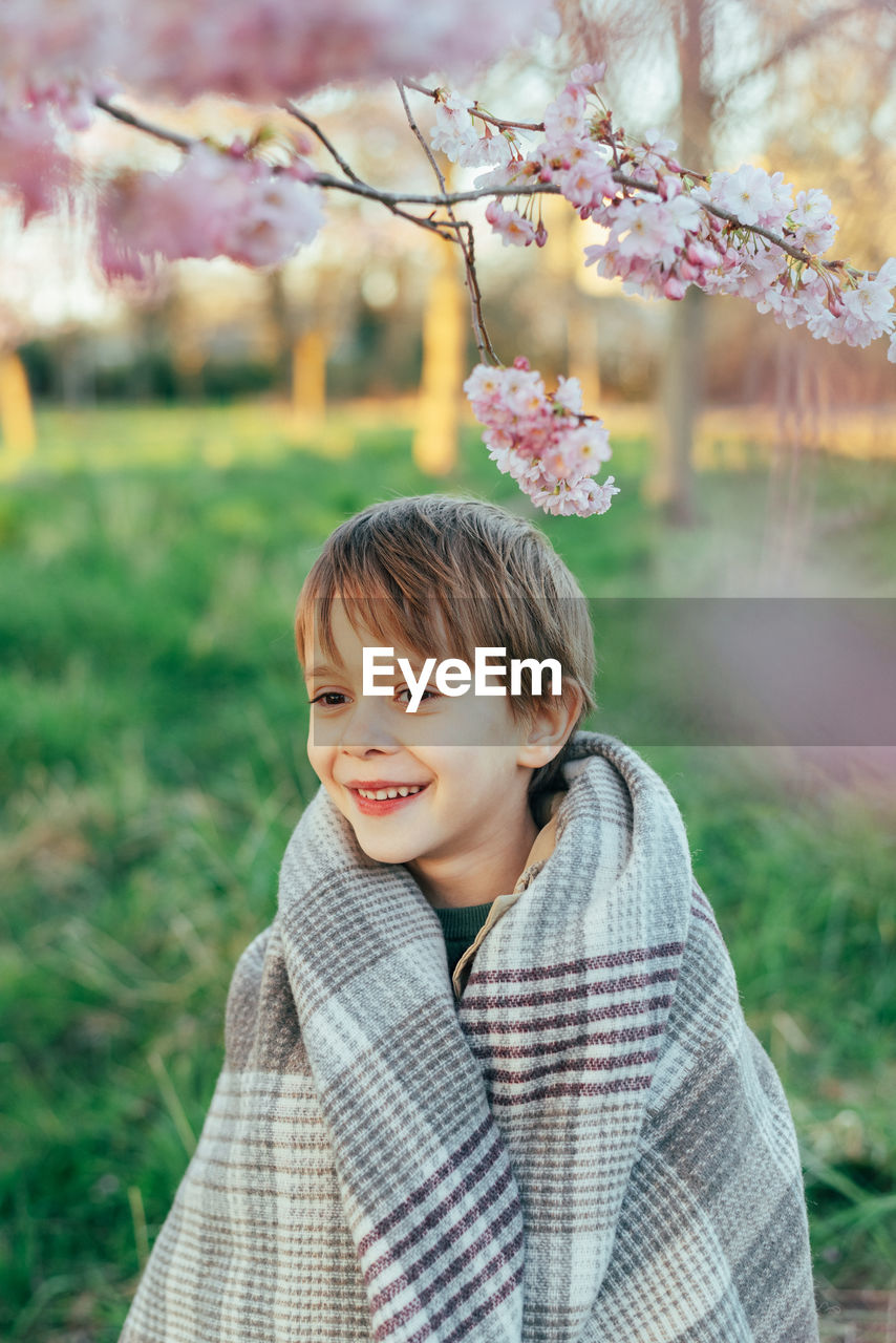 Portrait of a funny little boy wrapped in a blanket enjoying cherry blossoms in a city park.