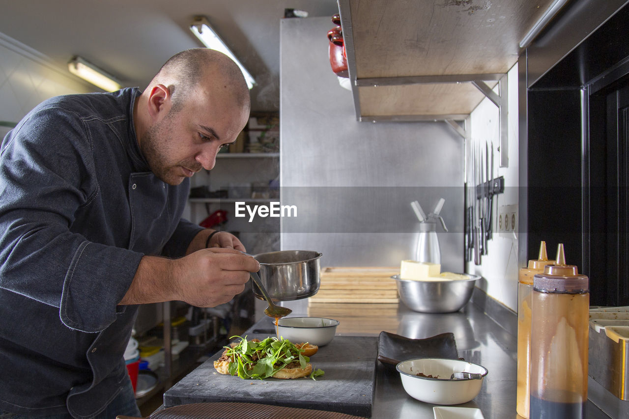 Side view of chef pouring tasty sauce on bun of sandwich with meat and greens on cutting board at counter in light kitchen