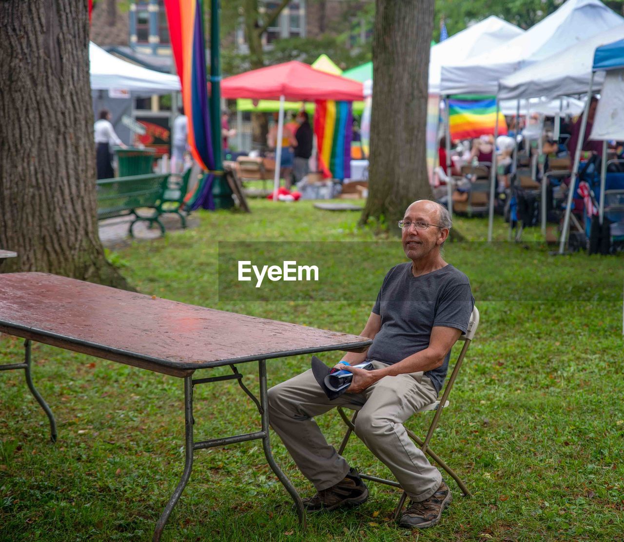 A smiling man sits alone at outdoor pride fest with rainbow flags