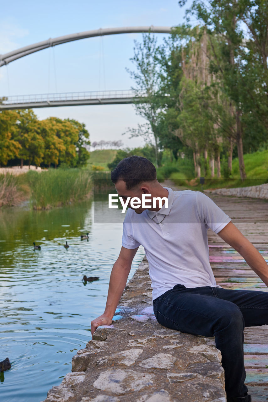 An attractive young man sitting on the bank of a canal in a park.