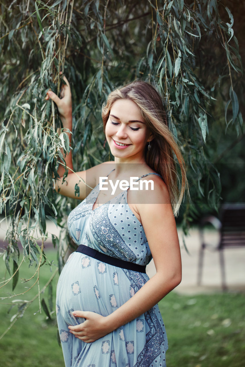 Smiling pregnant woman touching abdomen while standing on grass