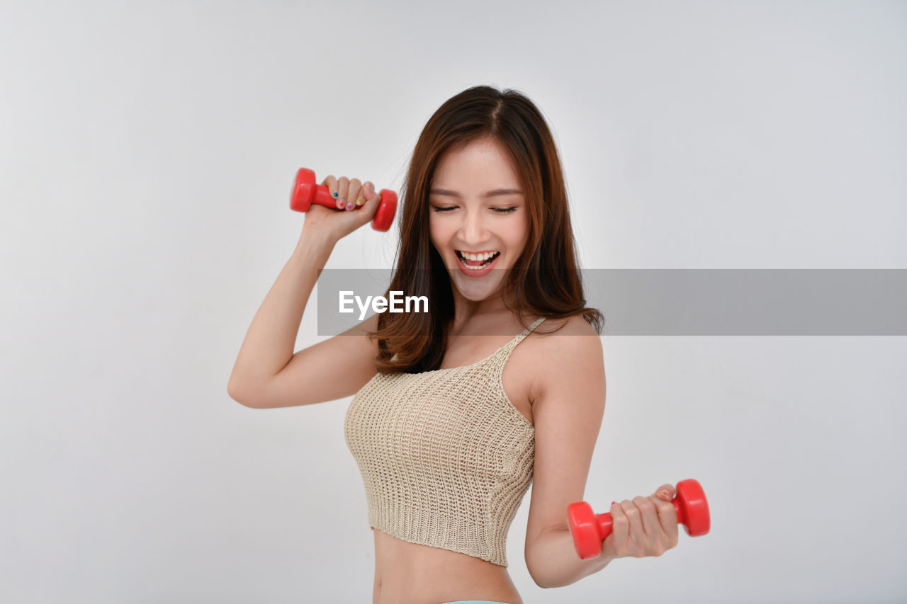 Smiling woman holding dumbbells while standing against wall