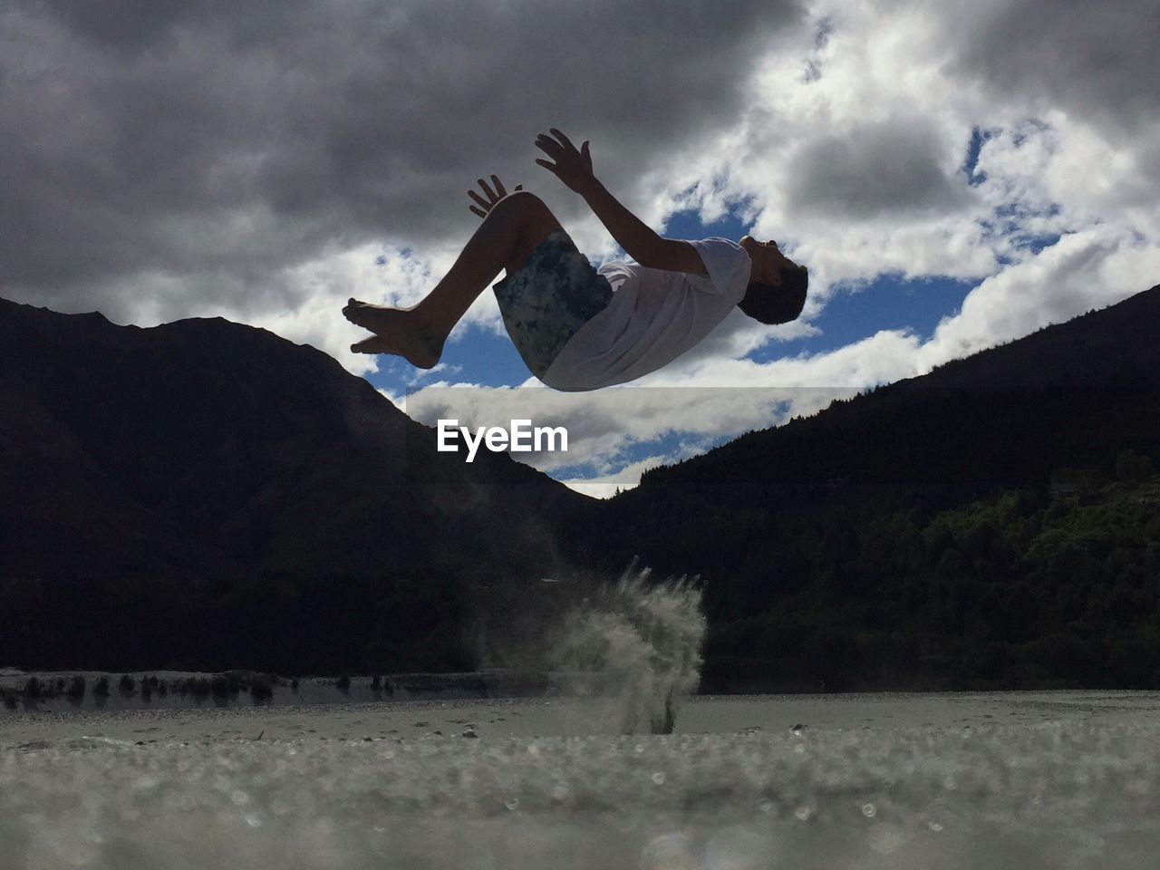 Full length of young man doing backflip over sand against cloudy sky