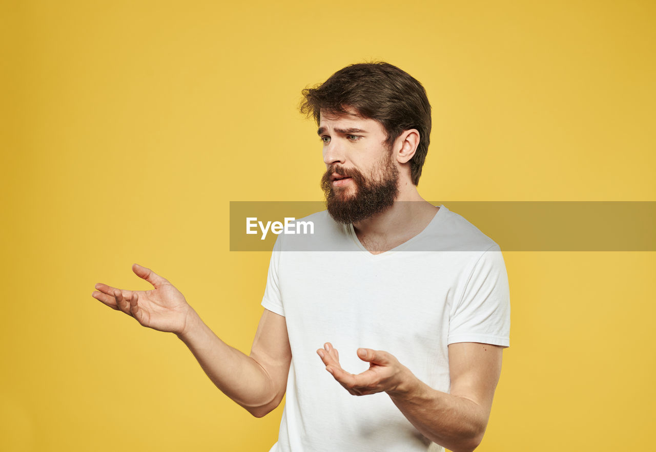 facial hair, colored background, beard, yellow, one person, adult, studio shot, yellow background, men, waist up, indoors, young adult, casual clothing, person, standing, copy space, t-shirt, smiling, portrait, emotion, looking, communication, happiness, holding, lifestyles, brown hair, gesturing, finger, clothing