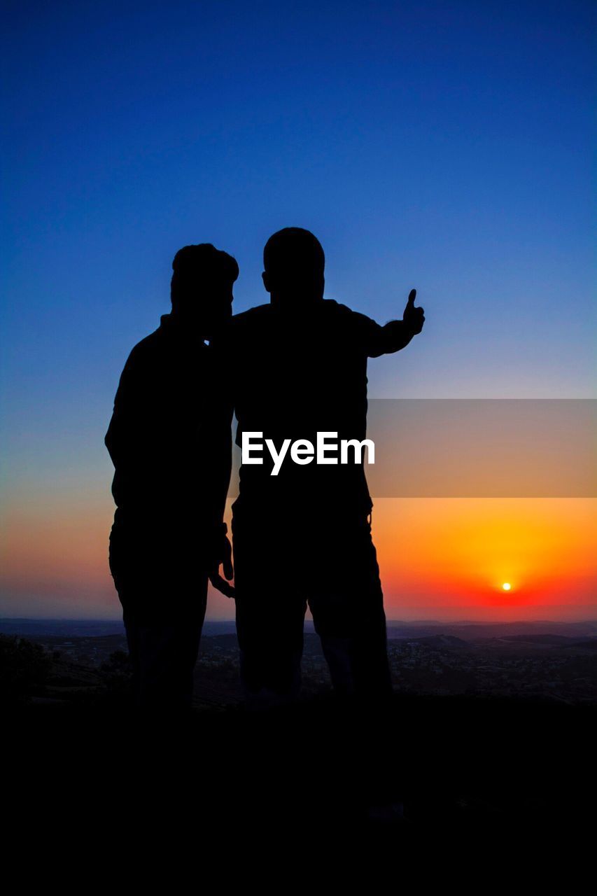 SILHOUETTE COUPLE STANDING ON BEACH AGAINST SKY DURING SUNSET