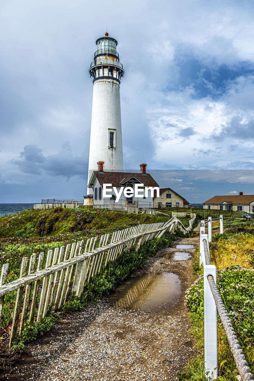 LIGHTHOUSE AMIDST PLANTS AND BUILDINGS AGAINST SKY