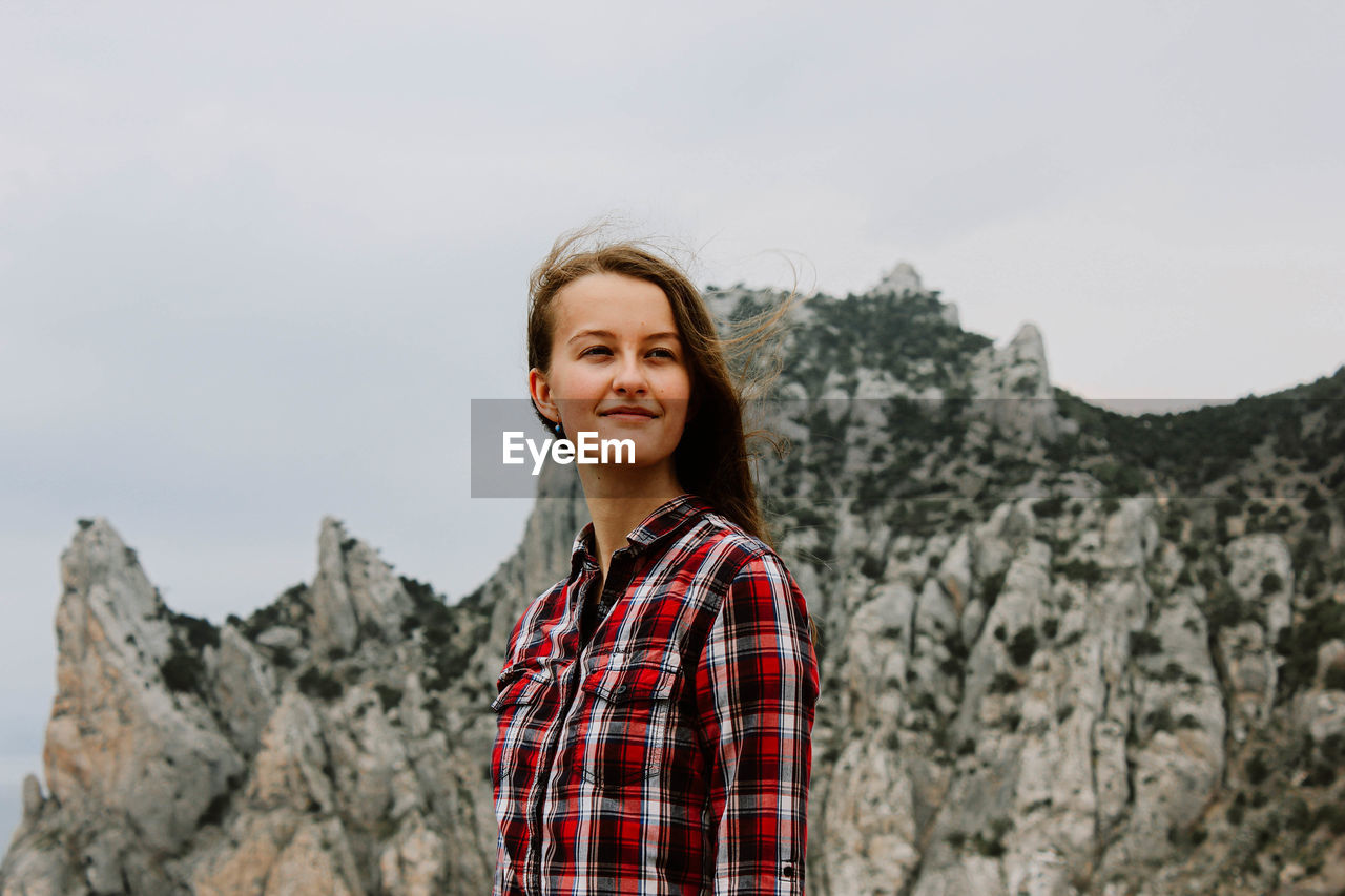 Portrait of smiling young woman standing on rock against sky