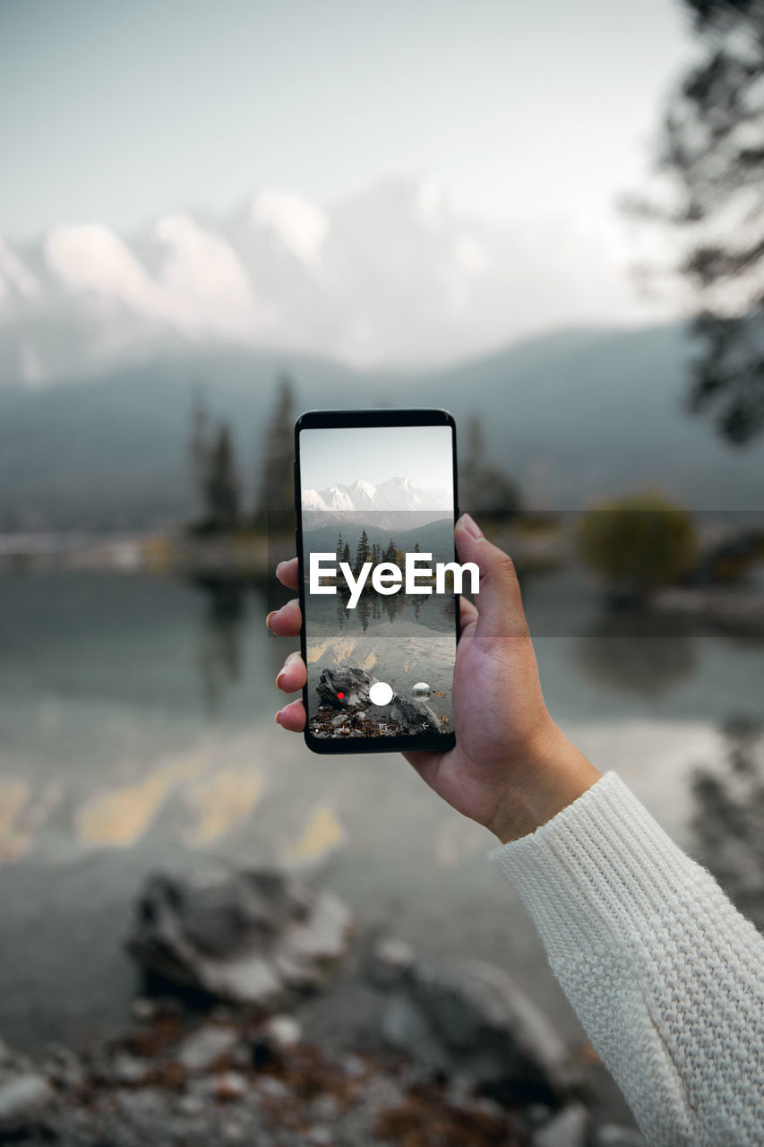 Cropped hand of woman photographing with mobile phone against lake