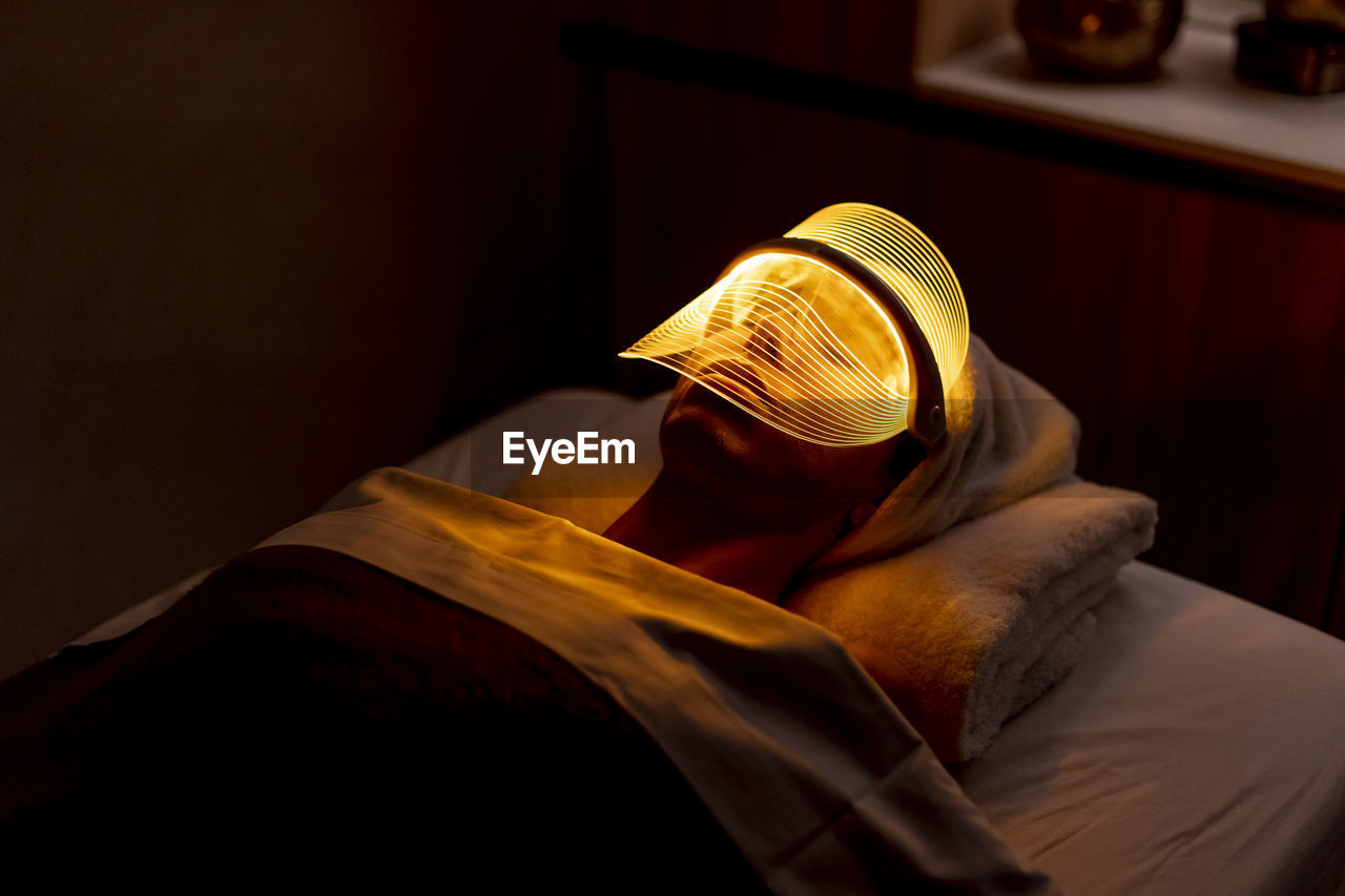 Young woman wearing yellow mask during rejuvenating treatment in dark at beauty spa