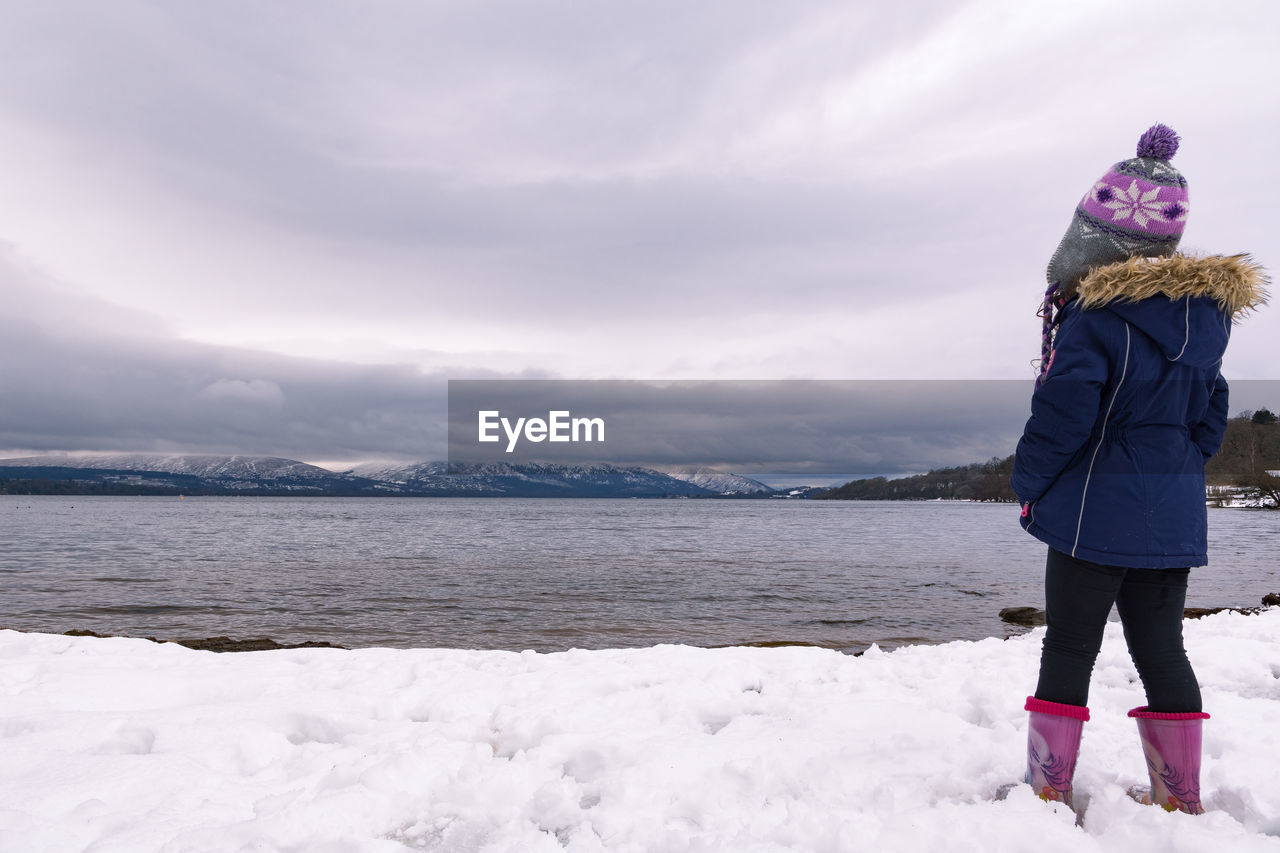 Rear view of girl standing at snow covered lakeshore against cloudy sky