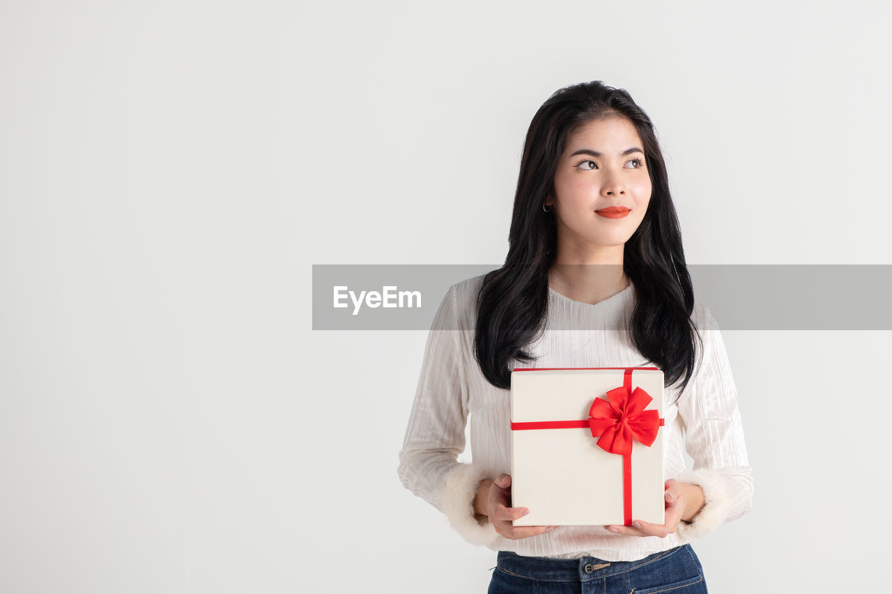 Beautiful young woman looking away holding gift standing against white background