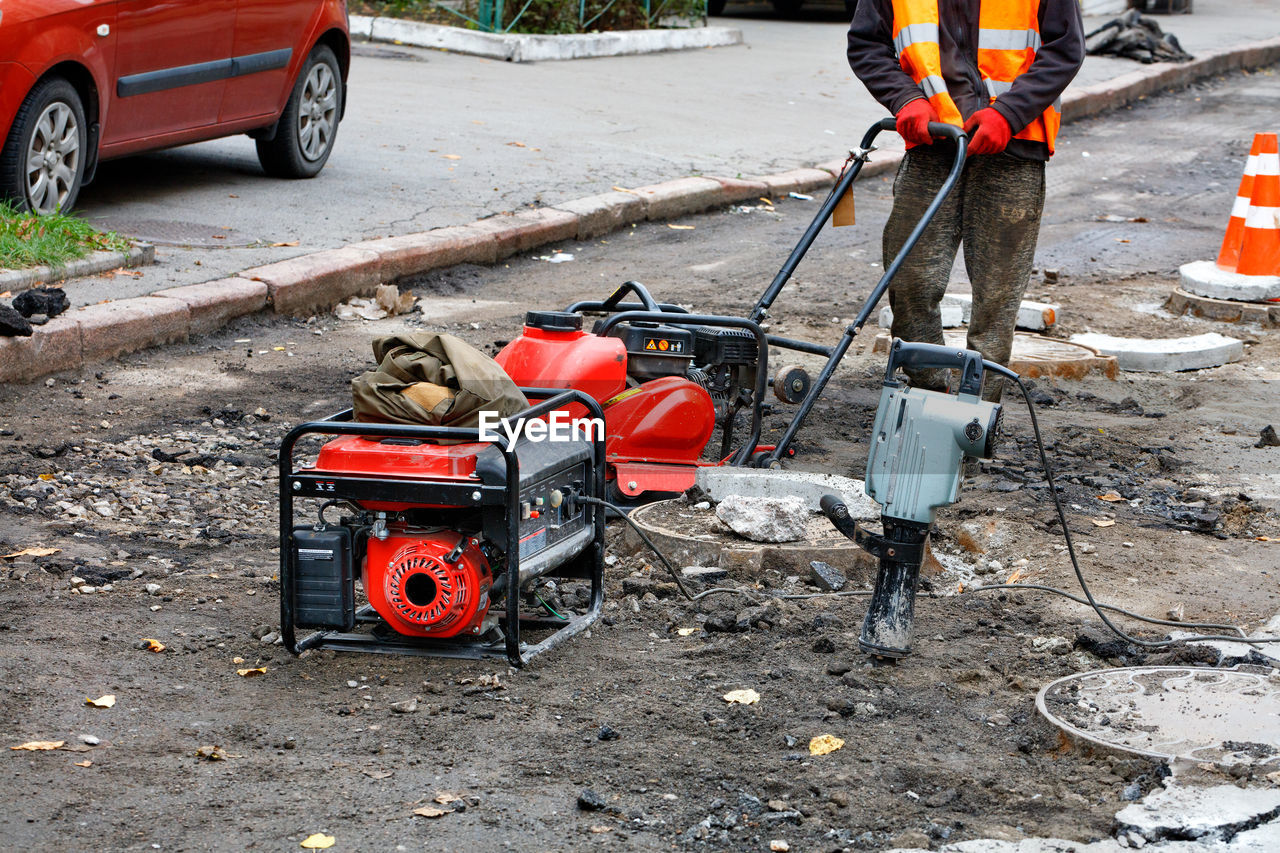 A road worker wearing a reflective orange vest repairs, installs sewers using compactor,  jackhammer