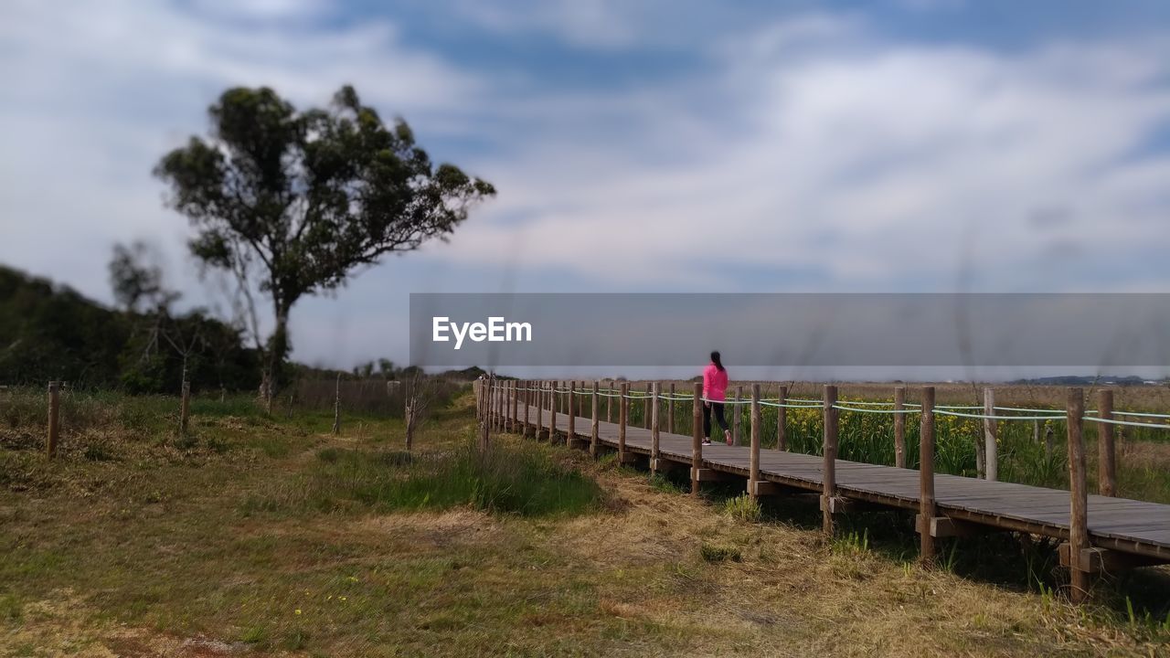 sky, plant, nature, cloud, landscape, one person, rural area, tree, land, environment, adult, full length, grass, walking, hill, fence, scenics - nature, outdoors, leisure activity, lifestyles, beauty in nature, women, railing, non-urban scene, tranquility, travel, day, field, waterway, rural scene, footpath, tranquil scene, architecture, transportation, bridge, standing