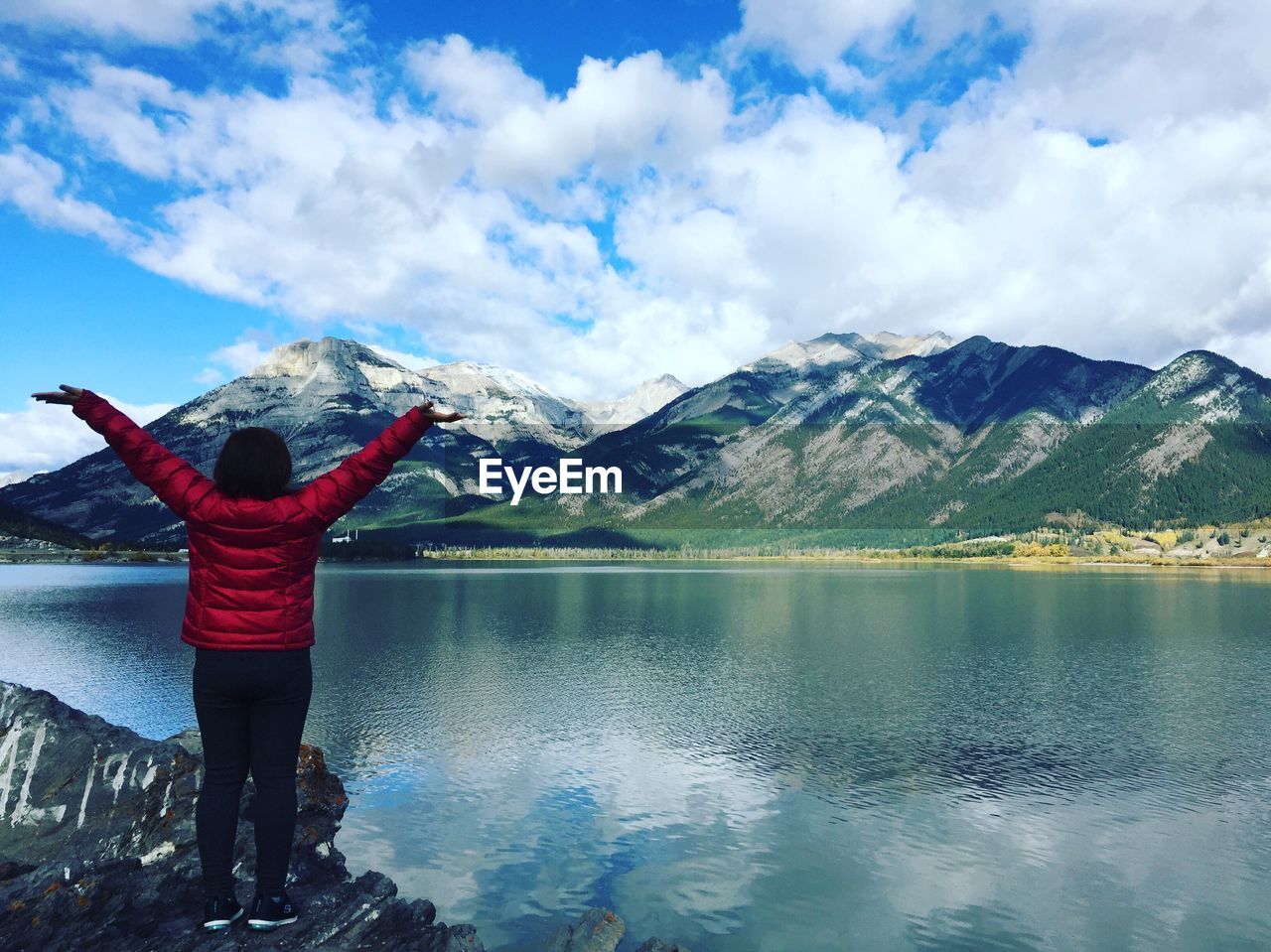 Rear view of woman with arms outstretched standing on rocks by lake and mountains against cloudy sky